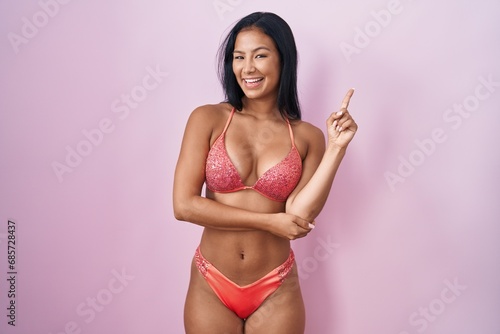 Hispanic woman wearing bikini with a big smile on face, pointing with hand finger to the side looking at the camera.