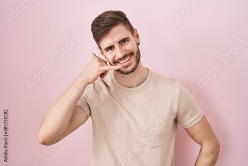 Hispanic man with beard standing over pink background smiling doing phone gesture with hand and fingers like talking on the telephone. communicating concepts.