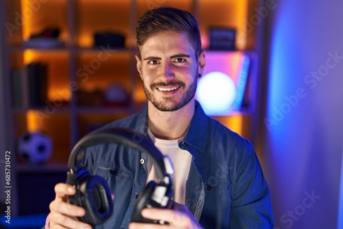 Young caucasian man streamer smiling confident holding headphones at gaming room