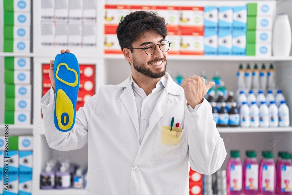 Hispanic man with beard working at pharmacy drugstore holding insole pointing thumb up to the side smiling happy with open mouth