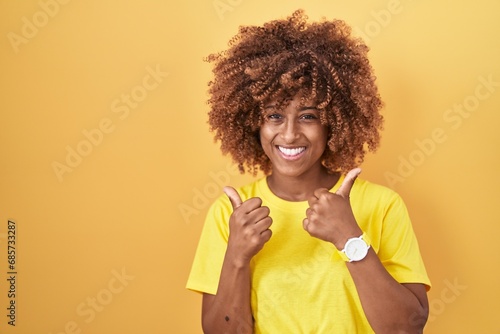 Young hispanic woman with curly hair standing over yellow background success sign doing positive gesture with hand, thumbs up smiling and happy. cheerful expression and winner gesture.
