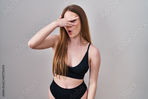 Young caucasian woman wearing lingerie peeking in shock covering face and eyes with hand, looking through fingers with embarrassed expression.