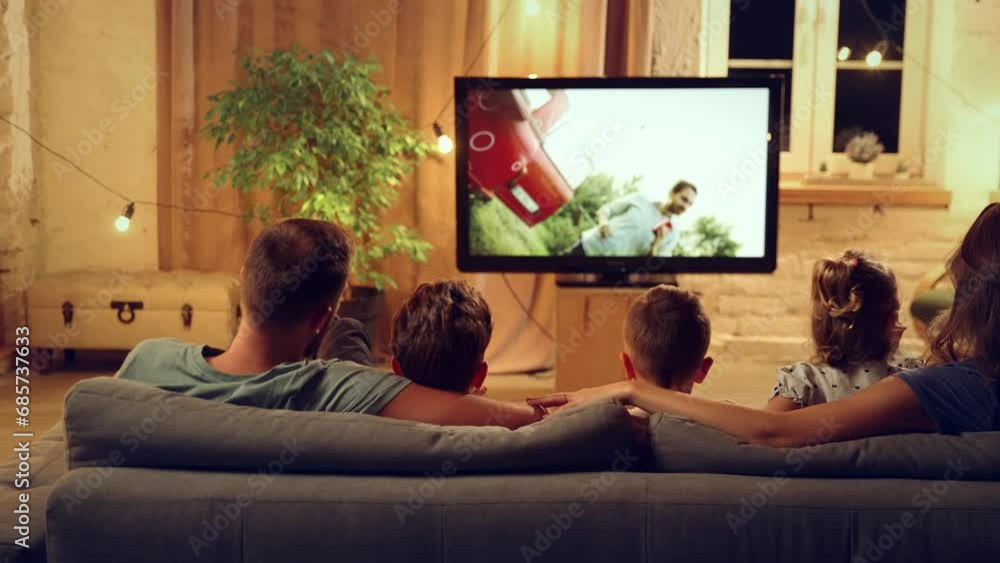 Back view of parents and children spending evening together, sitting on couch in living room and watching tv, eating snacks. Concept of family, leisure time, relaxation, childhood and parenthood Back view of parents and children spending evening together, sitting on couch in living room and watching tv, eating snacks. Concept of family, leisure time, relaxation, childhood and parenthood