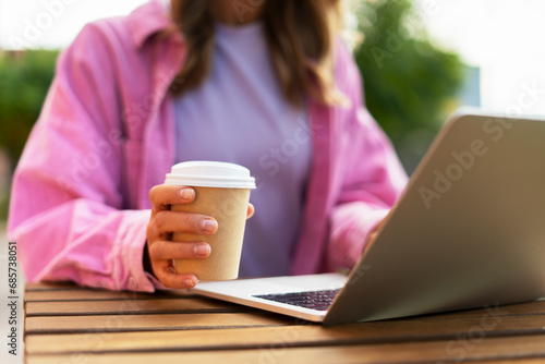 Closeup of student woman holding cup with coffee, sitting on street, using laptop