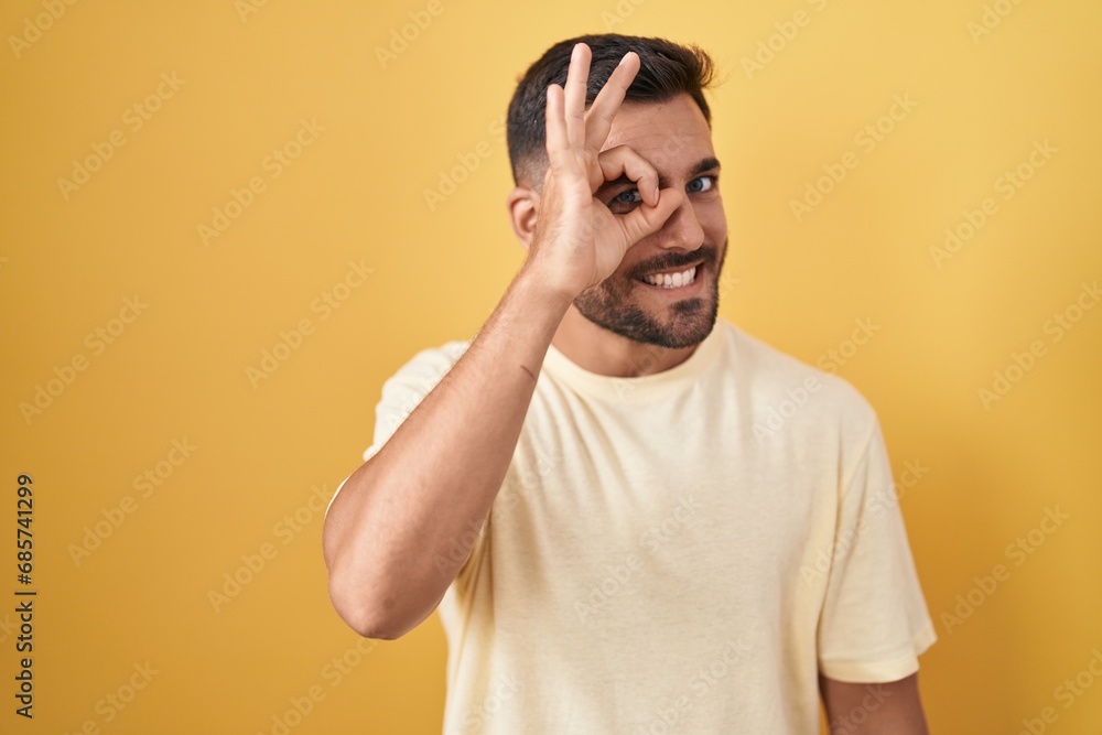 Handsome hispanic man standing over yellow background doing ok gesture with hand smiling, eye looking through fingers with happy face. Handsome hispanic man standing over yellow background doing ok gesture with hand smiling, eye looking through fingers with happy face.