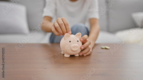 Adorable boy's hands inserting money into piggy bank while comfortably sitting on a living room sofa at home, symbolizing childhood financial learning