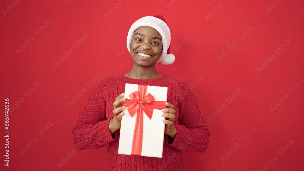 African american woman holding gift wearing christmas hat over isolated red background