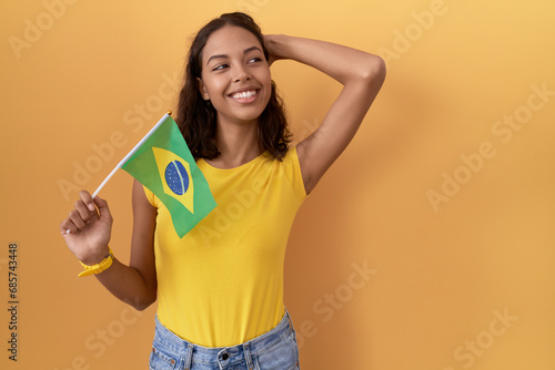Young hispanic woman holding brazil flag smiling confident touching hair with hand up gesture  posing attractive and fashionable
