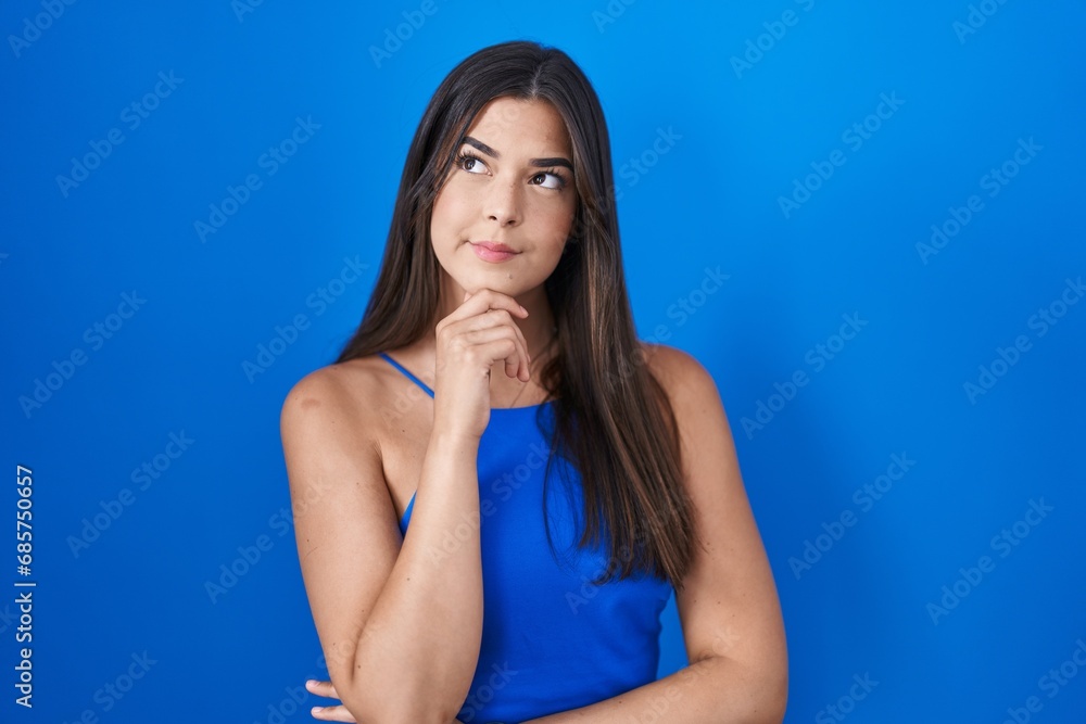 Hispanic woman standing over blue background with hand on chin thinking about question, pensive expression. smiling with thoughtful face. doubt concept.