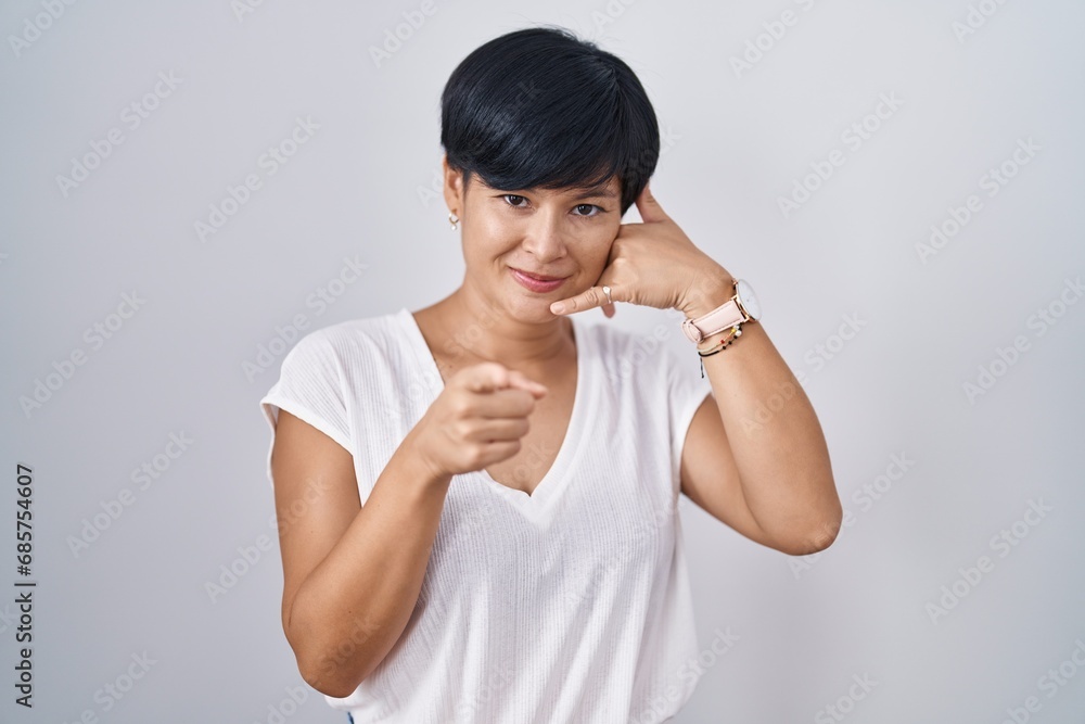 Young asian woman with short hair standing over isolated background smiling doing talking on the telephone gesture and pointing to you. call me. Young asian woman with short hair standing over isolated background smiling doing talking on the telephone gesture and pointing to you. call me.