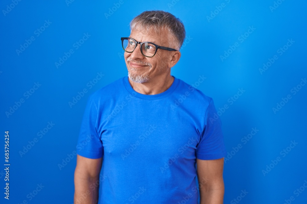 Hispanic man with grey hair standing over blue background smiling looking to the side and staring away thinking. - obrazy, fototapety, plakaty 
