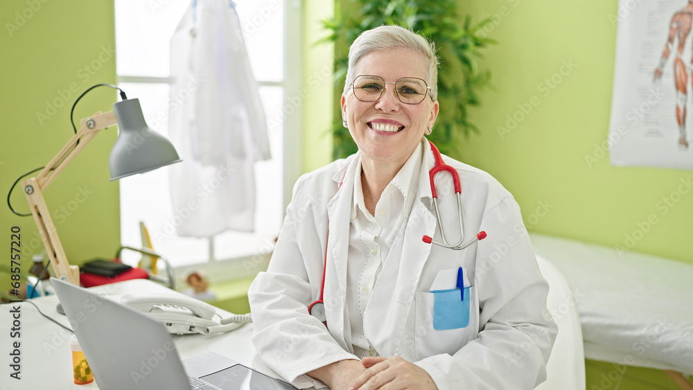 Middle age grey-haired woman doctor using laptop smiling at clinic Middle age grey-haired woman doctor using laptop smiling at clinic