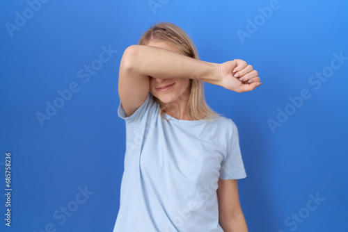 Young caucasian woman wearing casual blue t shirt covering eyes with arm smiling cheerful and funny. blind concept.