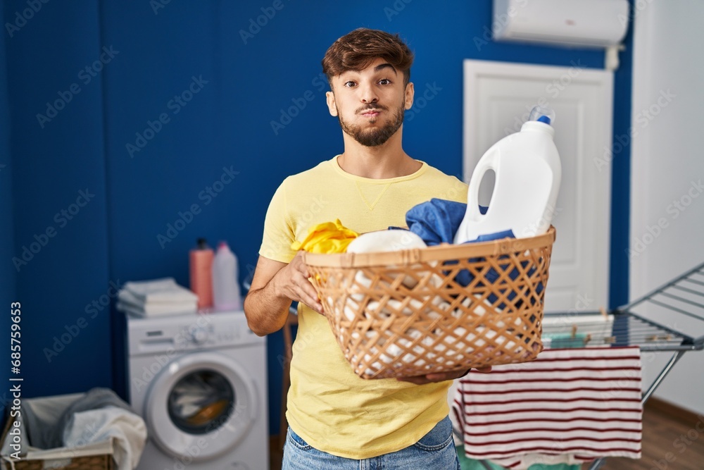 Arab man with beard holding laundry basket and detergent bottle puffing cheeks with funny face. mouth inflated with air, catching air. - obrazy, fototapety, plakaty 