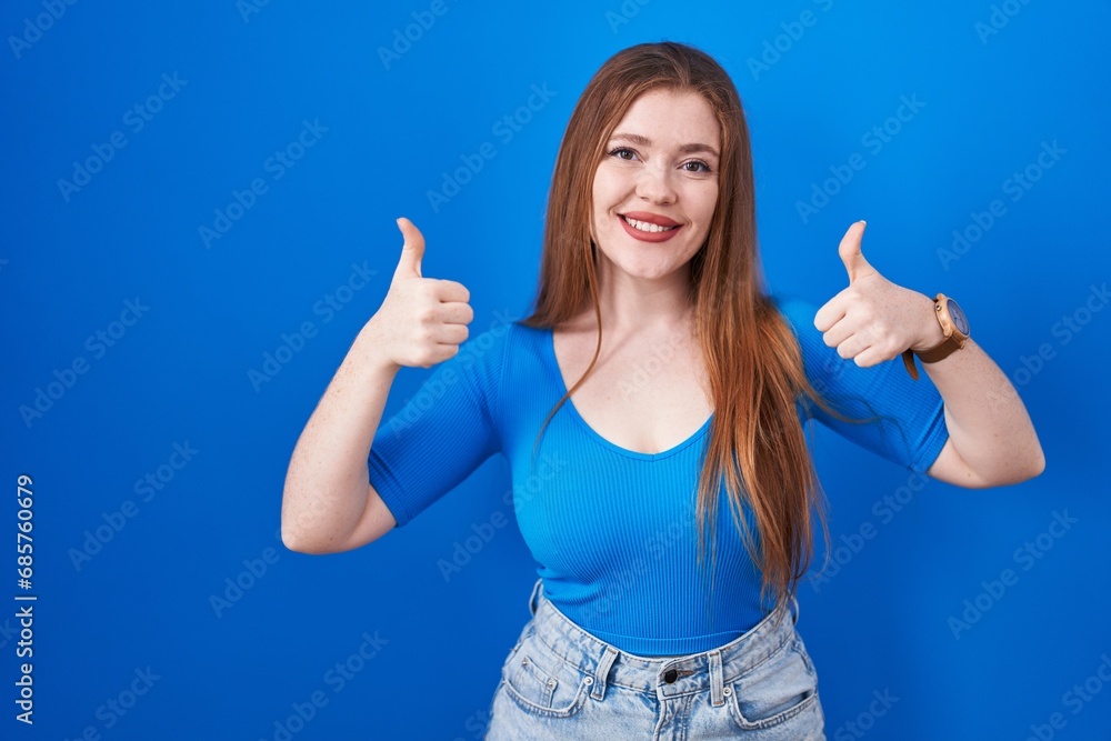 Redhead woman standing over blue background success sign doing positive gesture with hand, thumbs up smiling and happy. cheerful expression and winner gesture. Redhead woman standing over blue background success sign doing positive gesture with hand, thumbs up smiling and happy. cheerful expression and winner gesture.