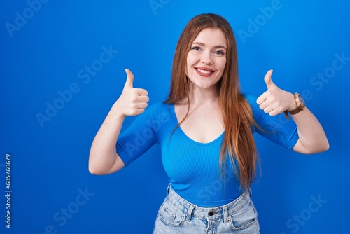 Redhead woman standing over blue background success sign doing positive gesture with hand, thumbs up smiling and happy. cheerful expression and winner gesture.