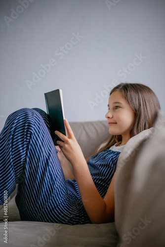 Vertical portrait of little girl sitting at home in relaxed position on sofa and carefully reading book