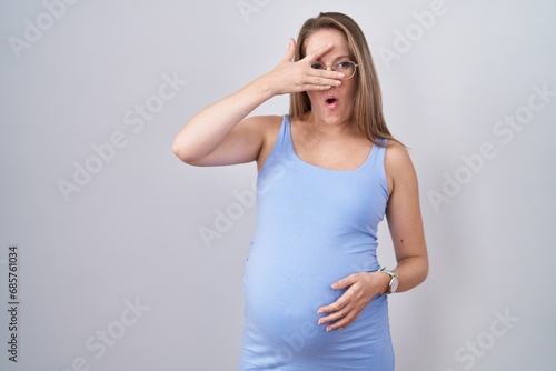 Young pregnant woman standing over white background peeking in shock covering face and eyes with hand  looking through fingers with embarrassed expression.