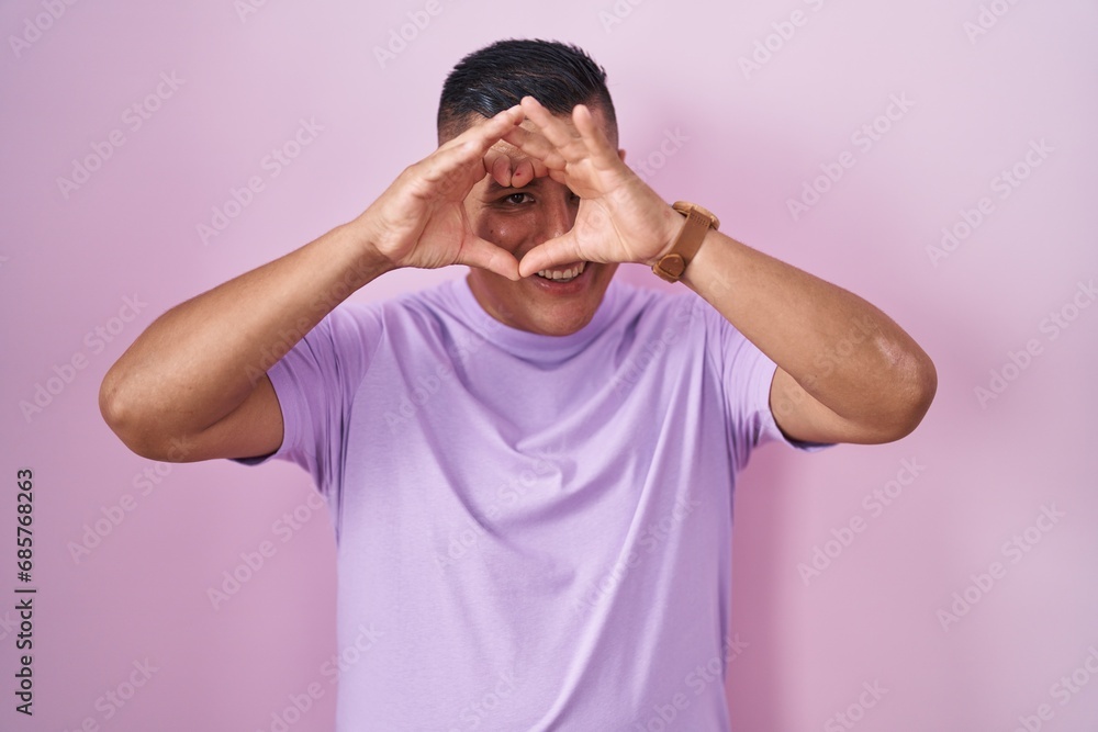 Young hispanic man standing over pink background doing heart shape with hand and fingers smiling looking through sign Young hispanic man standing over pink background doing heart shape with hand and fingers smiling looking through sign