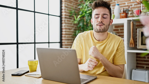 Young hispanic man student using laptop sitting on table dancing at dinning room