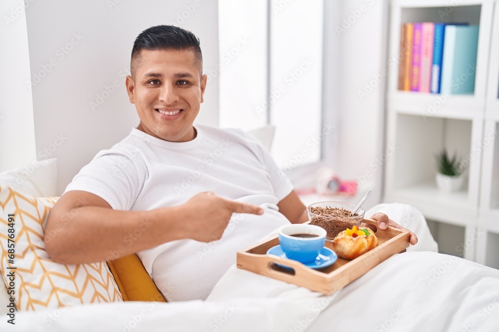 Young hispanic man eating breakfast in the bed smiling happy pointing with hand and finger Young hispanic man eating breakfast in the bed smiling happy pointing with hand and finger
