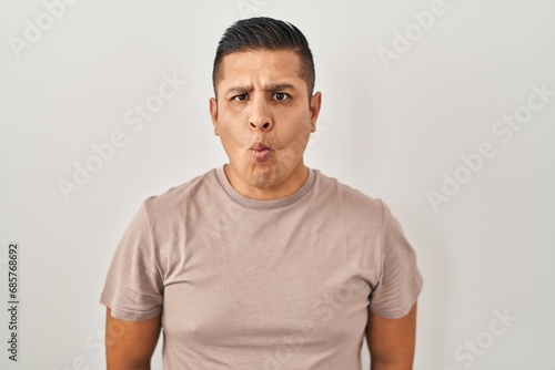 Hispanic young man standing over white background making fish face with lips, crazy and comical gesture. funny expression.