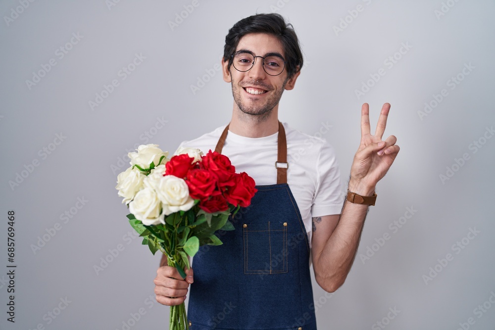 Young hispanic man holding bouquet of white and red roses smiling looking to the camera showing fingers doing victory sign. number two. Young hispanic man holding bouquet of white and red roses smiling looking to the camera showing fingers doing victory sign. number two.