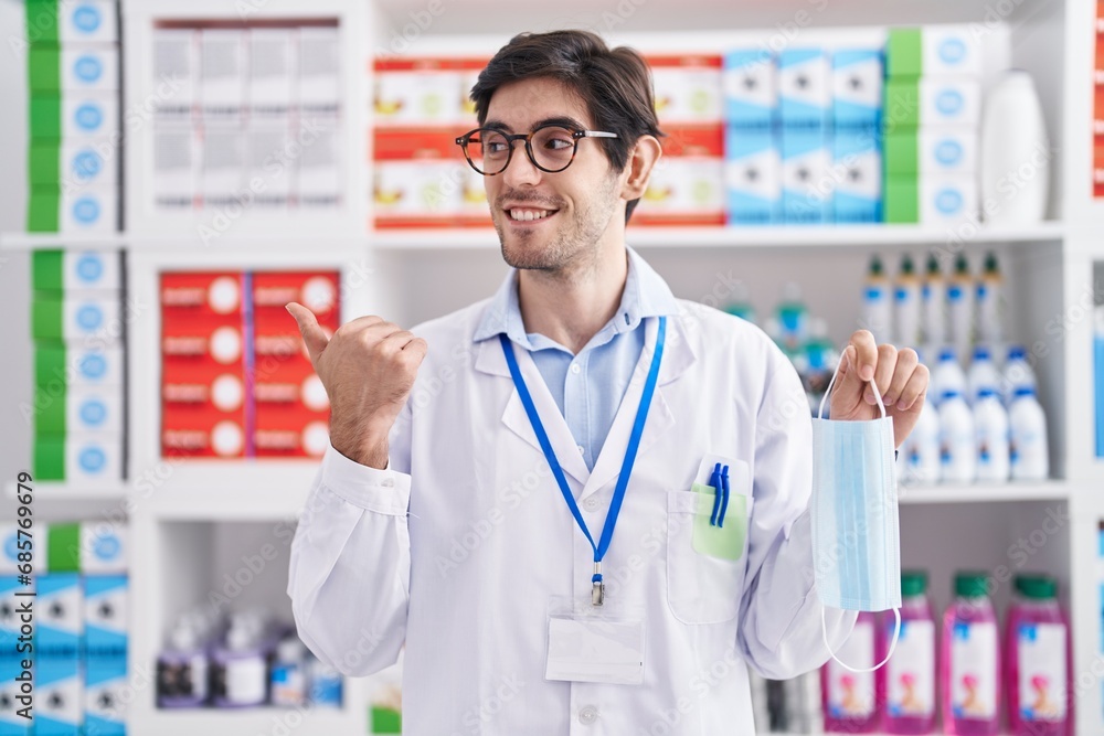 Young hispanic man working at pharmacy drugstore holding safety mask pointing thumb up to the side smiling happy with open mouth Young hispanic man working at pharmacy drugstore holding safety mask pointing thumb up to the side smiling happy with open mouth