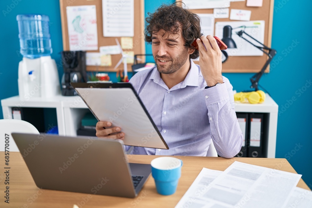 Young hispanic man business worker listening voice message by smartphone reading document at office