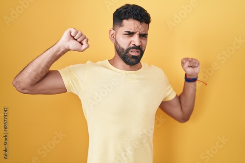 Hispanic man with beard standing over yellow background stretching back, tired and relaxed, sleepy and yawning for early morning