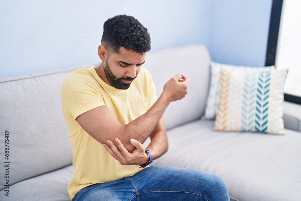 Young arab man sitting on sofa suffering for elbow pain at home Young arab man sitting on sofa suffering for elbow pain at home