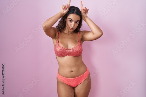 Young hispanic woman wearing lingerie over pink background doing funny gesture with finger over head as bull horns