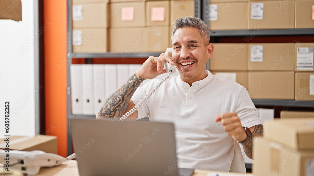 Young hispanic man ecommerce business worker using laptop talking on telephone celebrating at office Young hispanic man ecommerce business worker using laptop talking on telephone celebrating at office