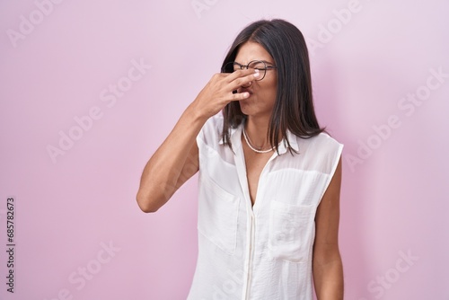 Brunette young woman standing over pink background wearing glasses smelling something stinky and disgusting, intolerable smell, holding breath with fingers on nose. bad smell