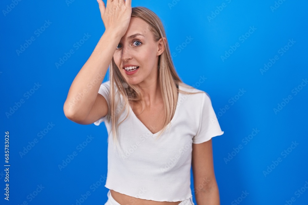 Fototapeta premium Young caucasian woman standing over blue background surprised with hand on head for mistake, remember error. forgot, bad memory concept.