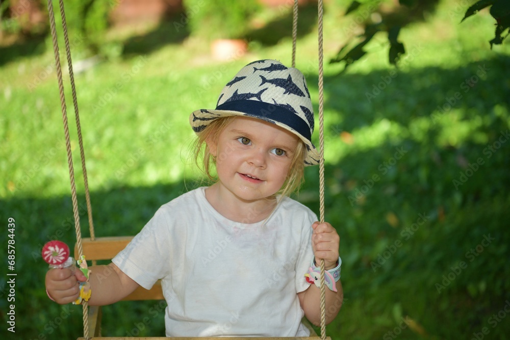 A little girl in a hat is swinging on a swing hung on a tree in the garden