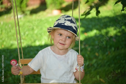 Obraz na płótnie A little girl in a hat is swinging on a swing hung on a tree in the garden w salonie