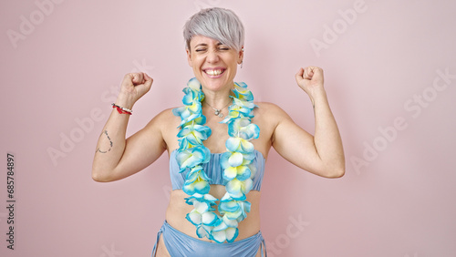 Young woman tourist wearing bikini and hawaiian lei with winner gesture over isolated pink background