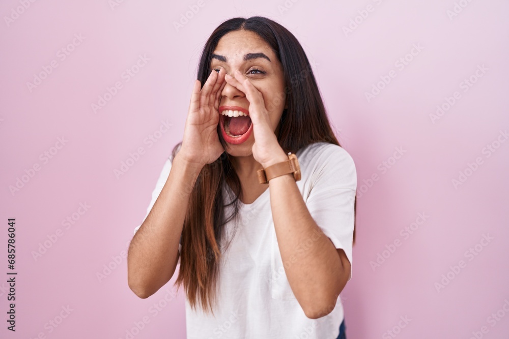 Young arab woman standing over pink background shouting angry out loud with hands over mouth