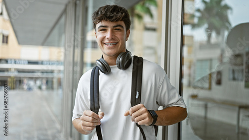 Cheerful young hispanic male student standing outdoors enjoying campus life, confidently wearing headphones and backpack, radiating positive energy and joy at the university.