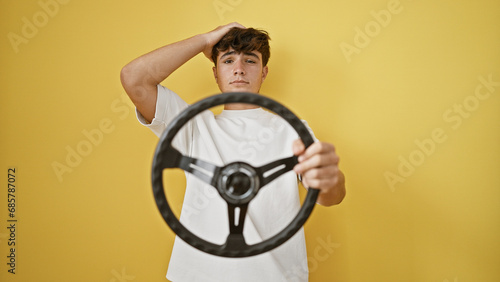 Young hispanic teenager, a serious yet upset novice driver, struggling with steering wheel against isolated yellow wall background