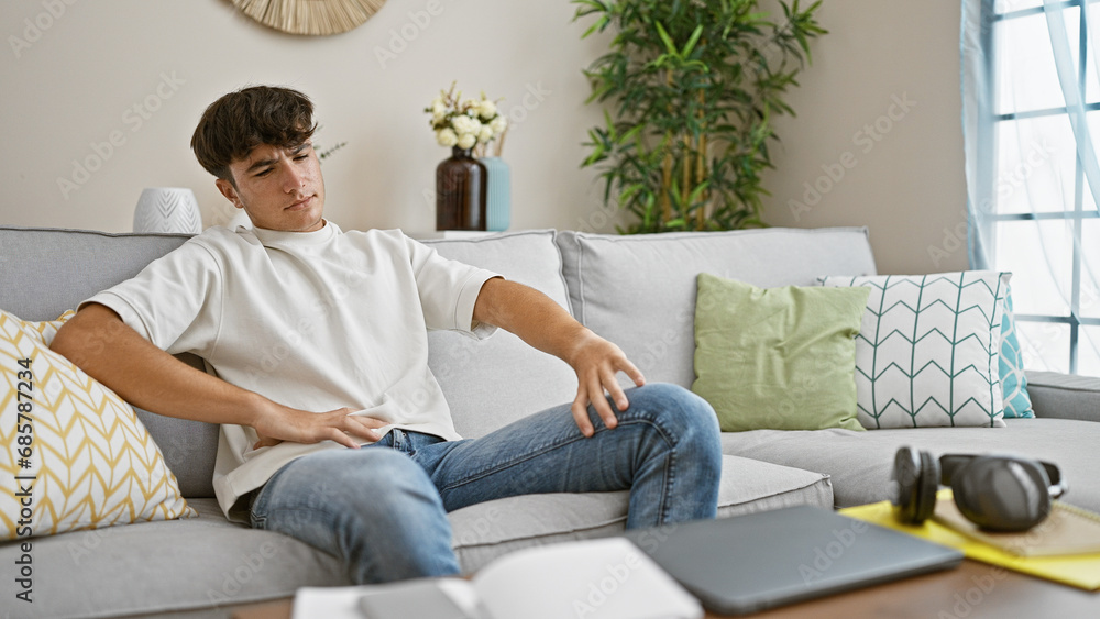 In the cozy comfort of his home, a young hispanic teenager is sitting on the sofa, a hint of doubt on his handsome face, seriously contemplating his thoughts while gazing off to the side. In the cozy comfort of his home, a young hispanic teenager is sitting on the sofa, a hint of doubt on his handsome face, seriously contemplating his thoughts while gazing off to the side.