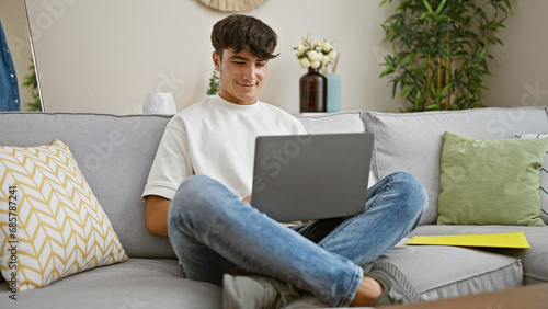 Konstfotografi Handsome young hispanic teenager beams with joy, comfortably sitting on sofa, using laptop confidently at home, embodying happiness & relaxation in living room's warm interior