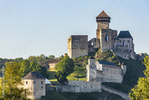 Trenčín castle with walls standing on a hill, surrounded by greenery and illuminated by the rising sun.