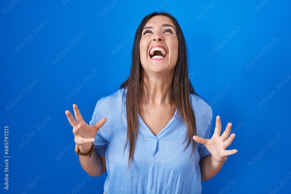 Young brunette woman standing over blue background angry and mad screaming frustrated and furious, shouting with anger. rage and aggressive concept. Young brunette woman standing over blue background angry and mad screaming frustrated and furious, shouting with anger. rage and aggressive concept.