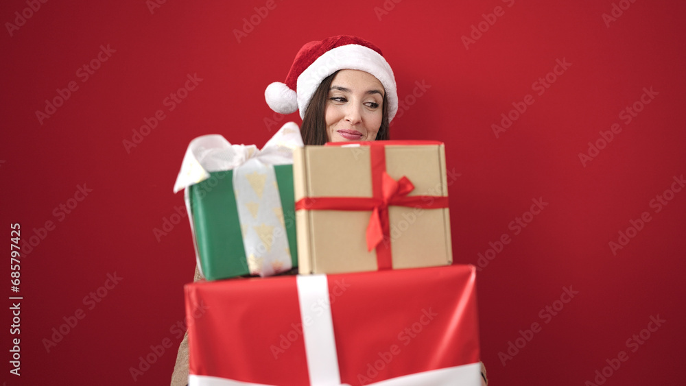 Beautiful hispanic woman wearing christmas hat holding gifts over isolated red background Beautiful hispanic woman wearing christmas hat holding gifts over isolated red background
