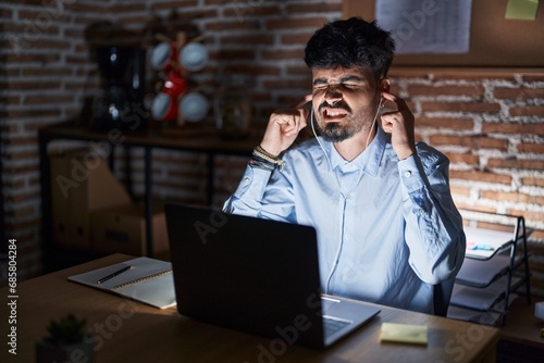 Young hispanic man with beard working at the office at night covering ears with fingers with annoyed expression for the noise of loud music. deaf concept.