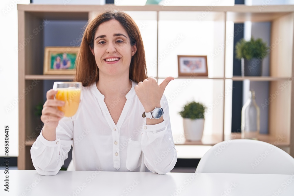 Brunette woman drinking glass of orange juice pointing to the back behind with hand and thumbs up, smiling confident Brunette woman drinking glass of orange juice pointing to the back behind with hand and thumbs up, smiling confident