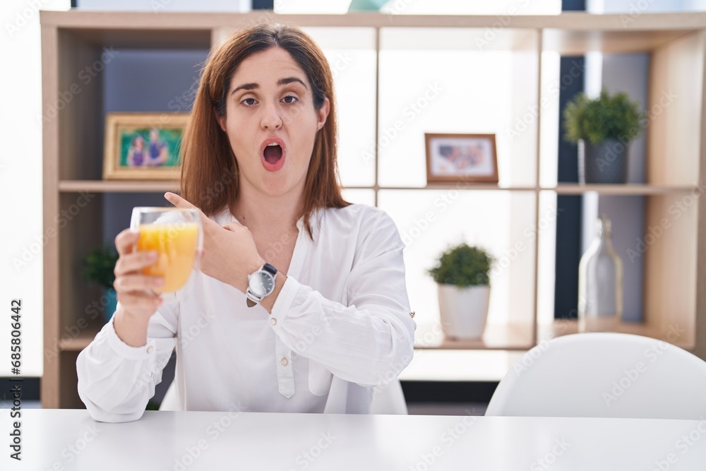 Brunette woman drinking glass of orange juice surprised pointing with finger to the side, open mouth amazed expression. Brunette woman drinking glass of orange juice surprised pointing with finger to the side, open mouth amazed expression.