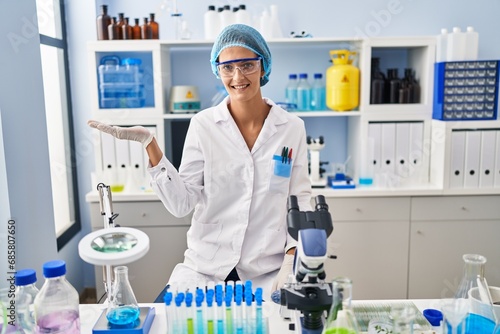 Brunette woman working at scientist laboratory smiling cheerful presenting and pointing with palm of hand looking at the camera.